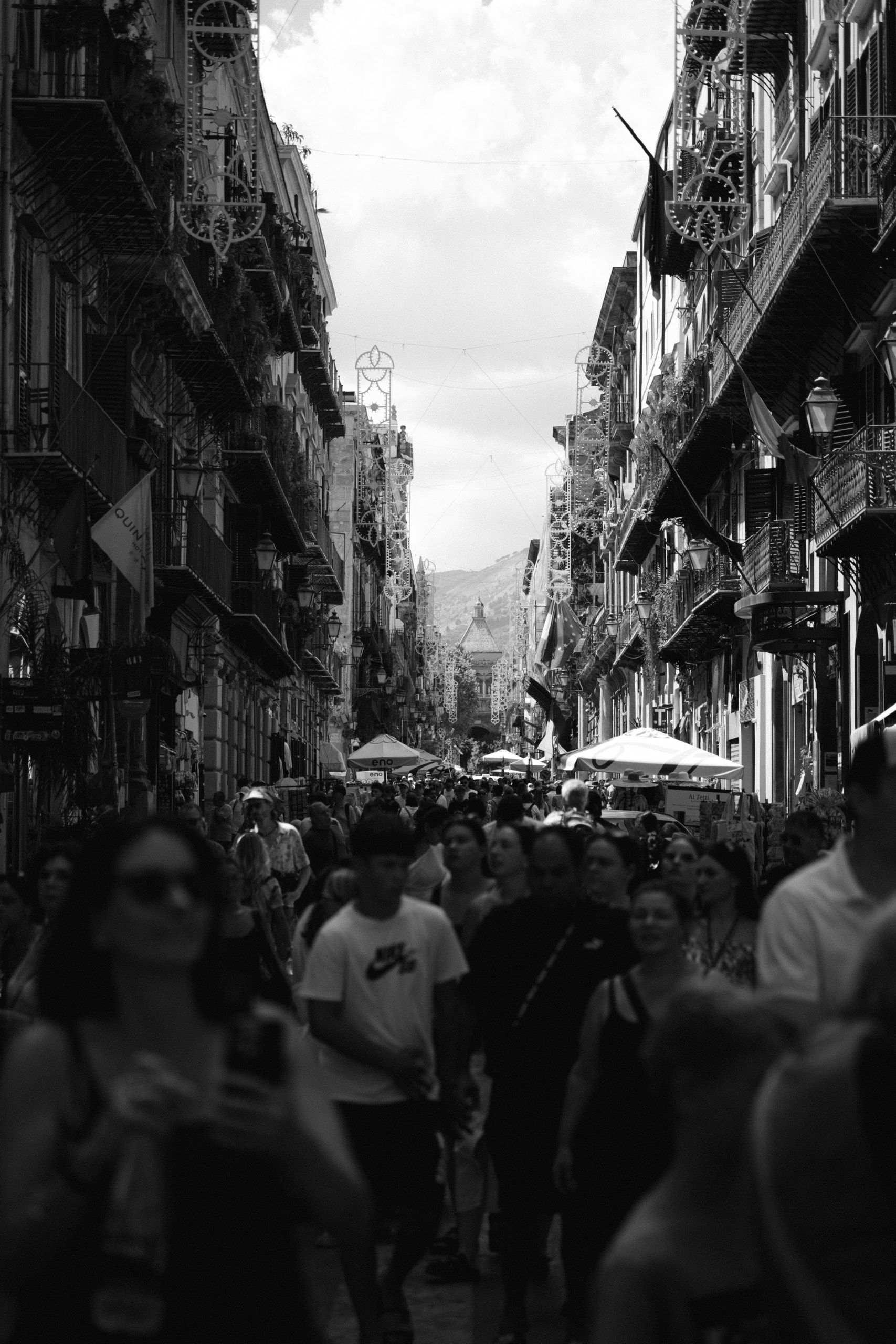 Una folla cammina in una lunga strada stretta tra palazzi con balconi, verso il fondo pieno di luce