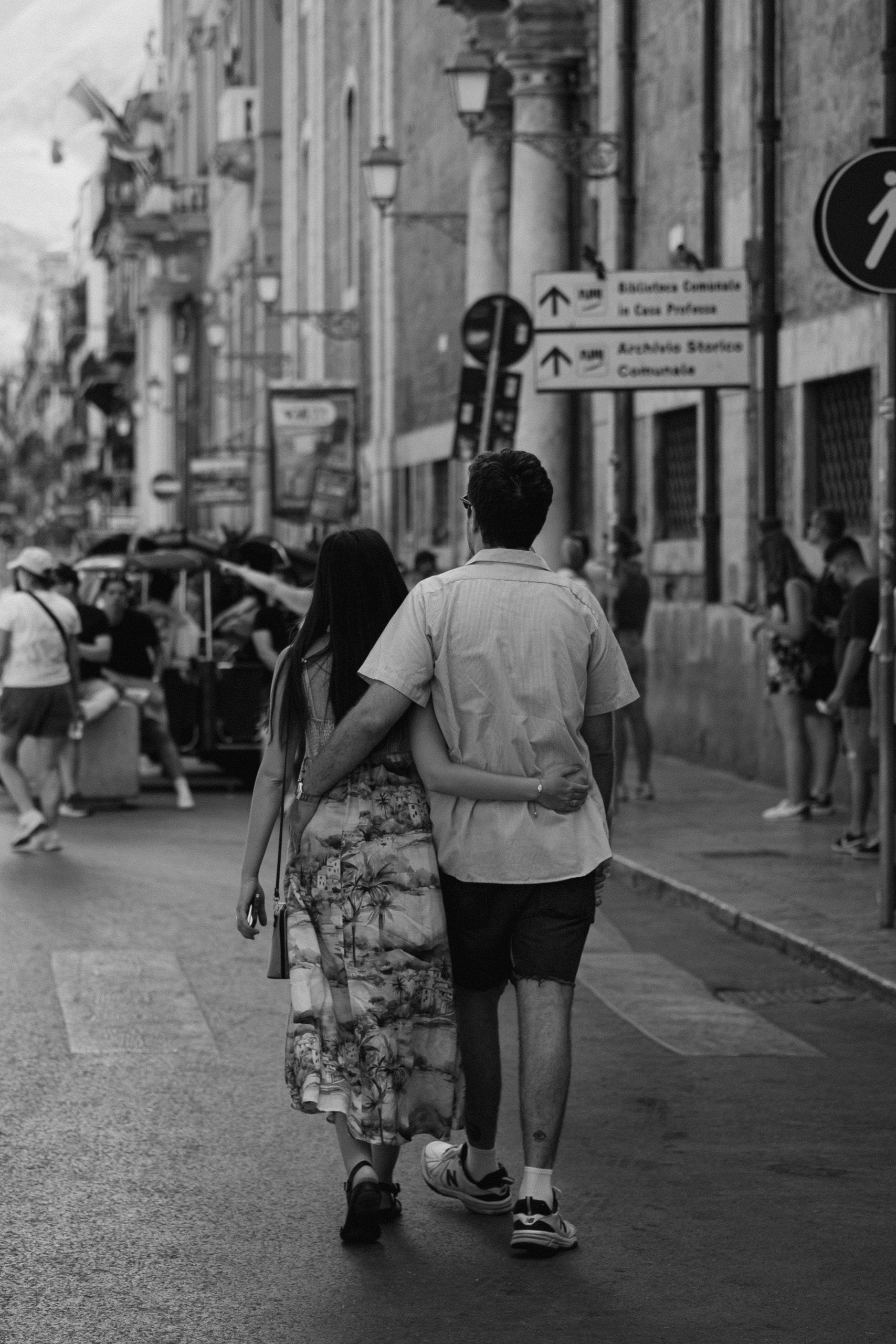 Una coppia di spalle cammina abbracciata in una strada affollata, tra palazzi antichi e cartelli stradali