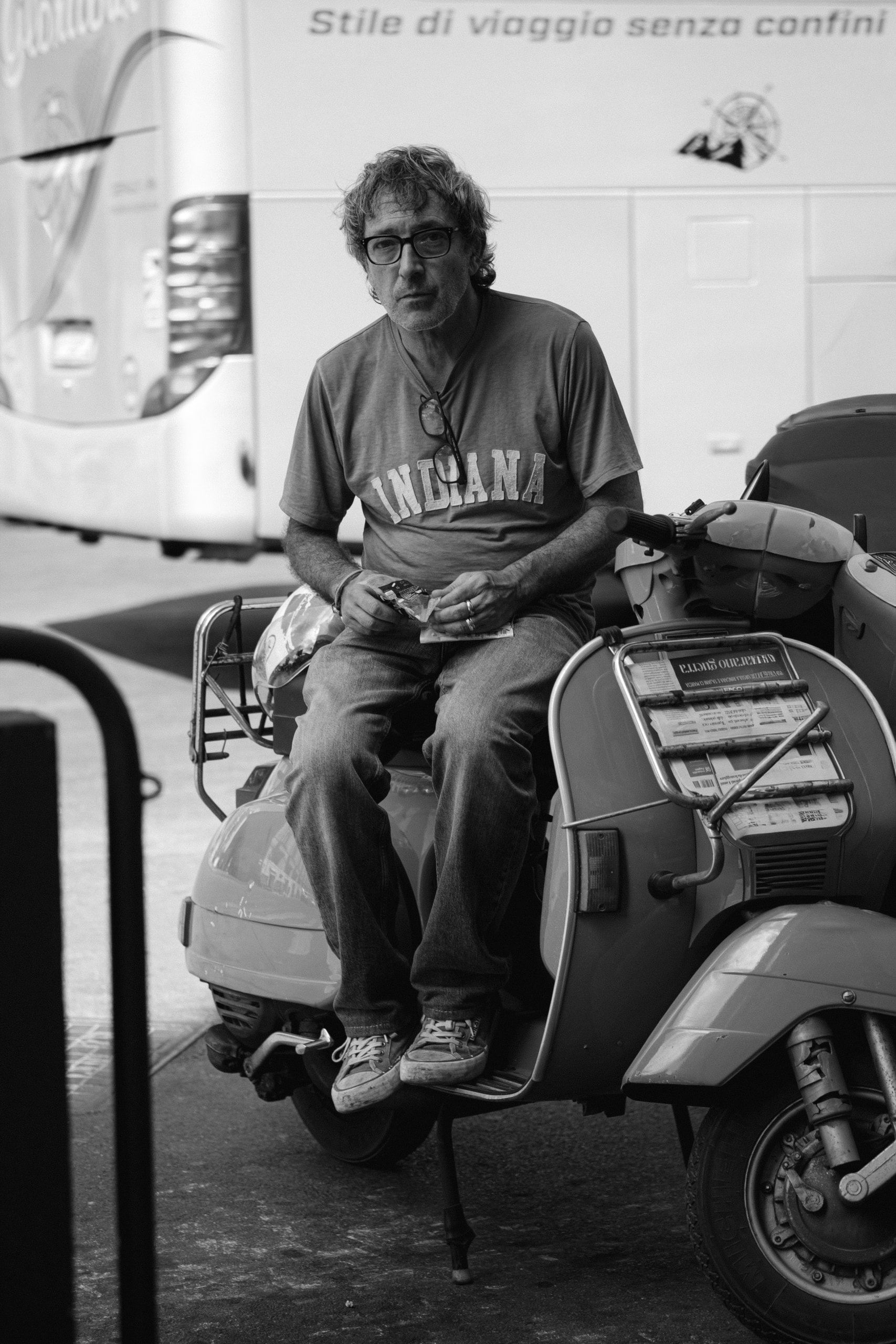 Un uomo con gli occhiali siede su uno scooter d'epoca e guarda l'obiettivo, con un pullman alle spalle