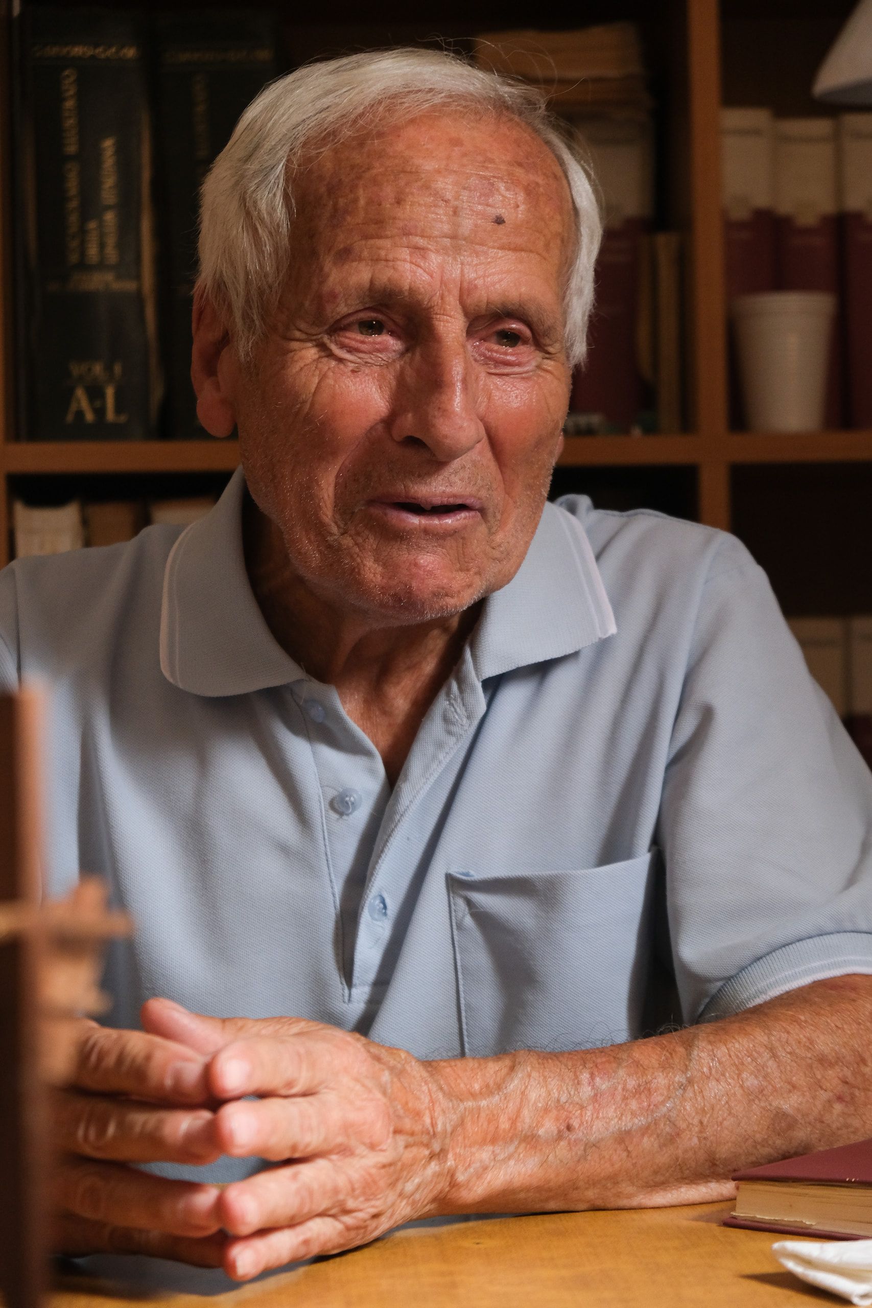 Filippo Castro parla con le mani raccolte sul tavolo, davanti alla libreria