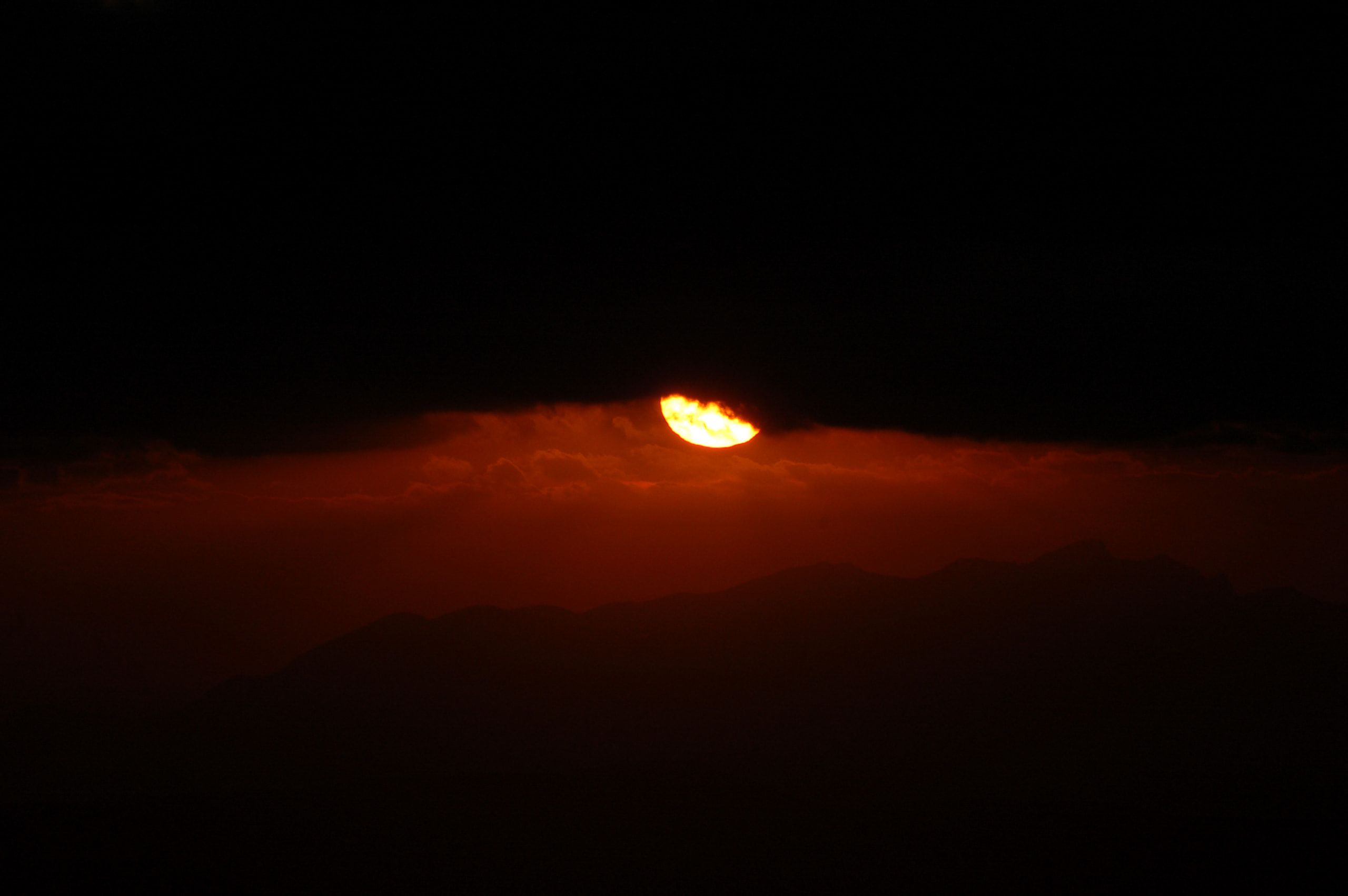 Il sole, nascosto per metà da una fascia di nuvole scure, sopra il profilo dei monti in un cielo rosso cupo