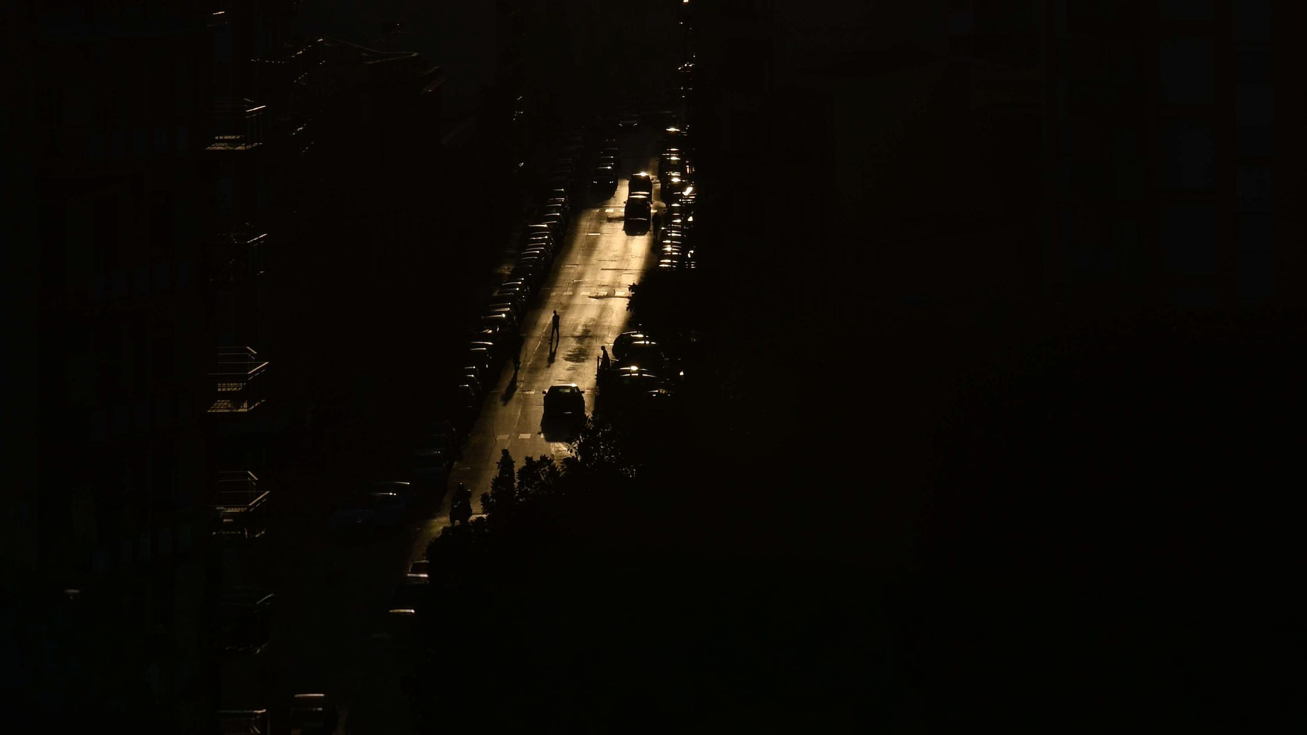 Una strada vista dall'alto, illuminata da una luce radente tra i palazzi in ombra, con auto e passanti in controluce
