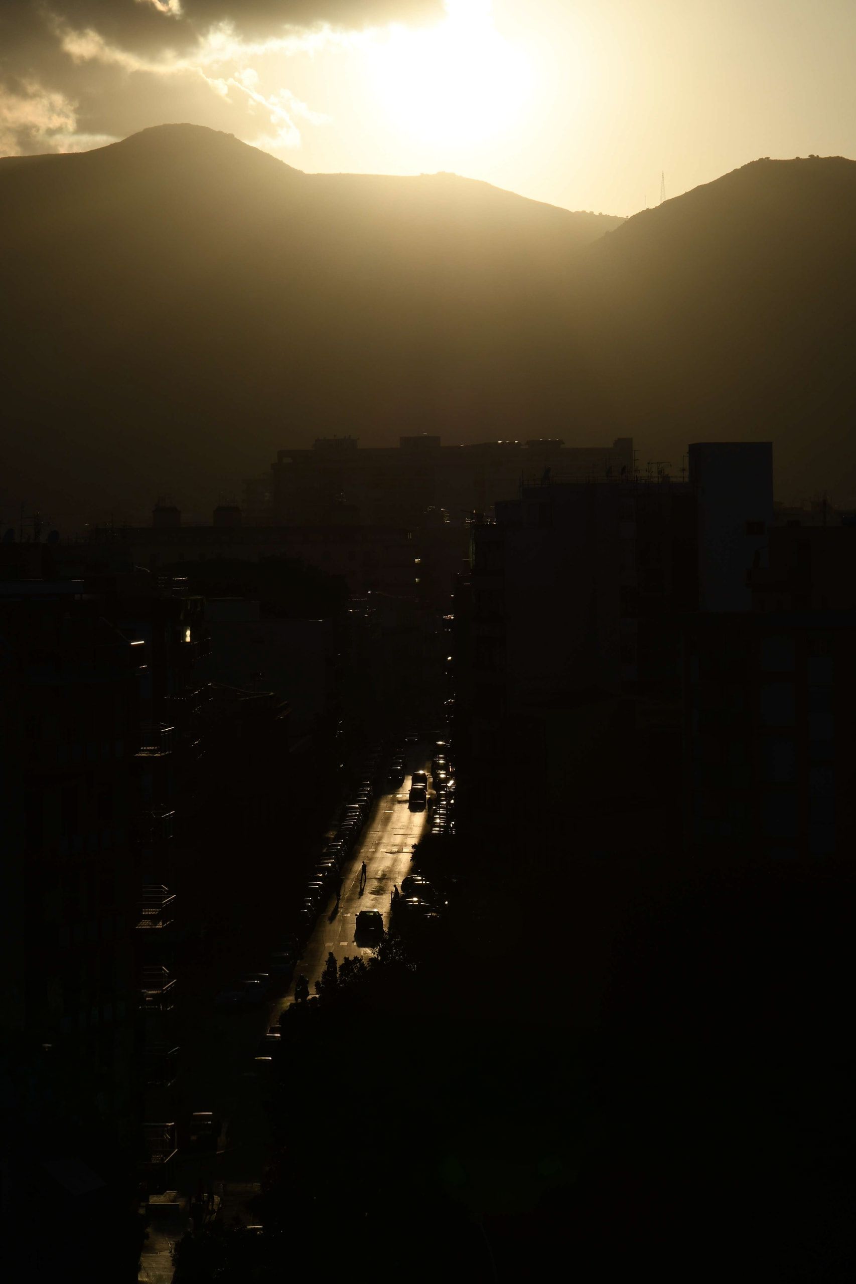 Una strada vista dall'alto tra palazzi in ombra, illuminata dal sole basso sopra i monti, con auto e passanti