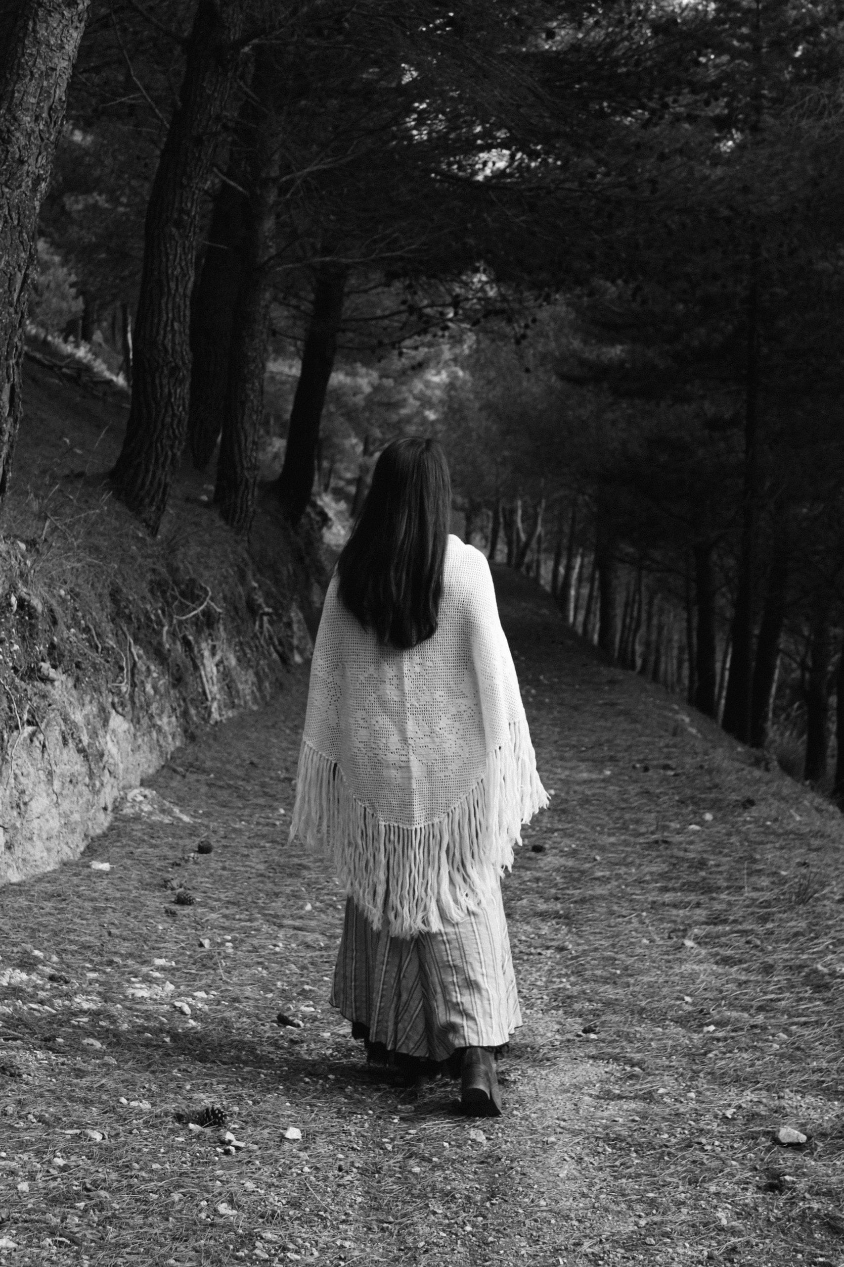 Una donna di spalle, con uno scialle a frange e la gonna lunga, cammina su un sentiero tra i pini