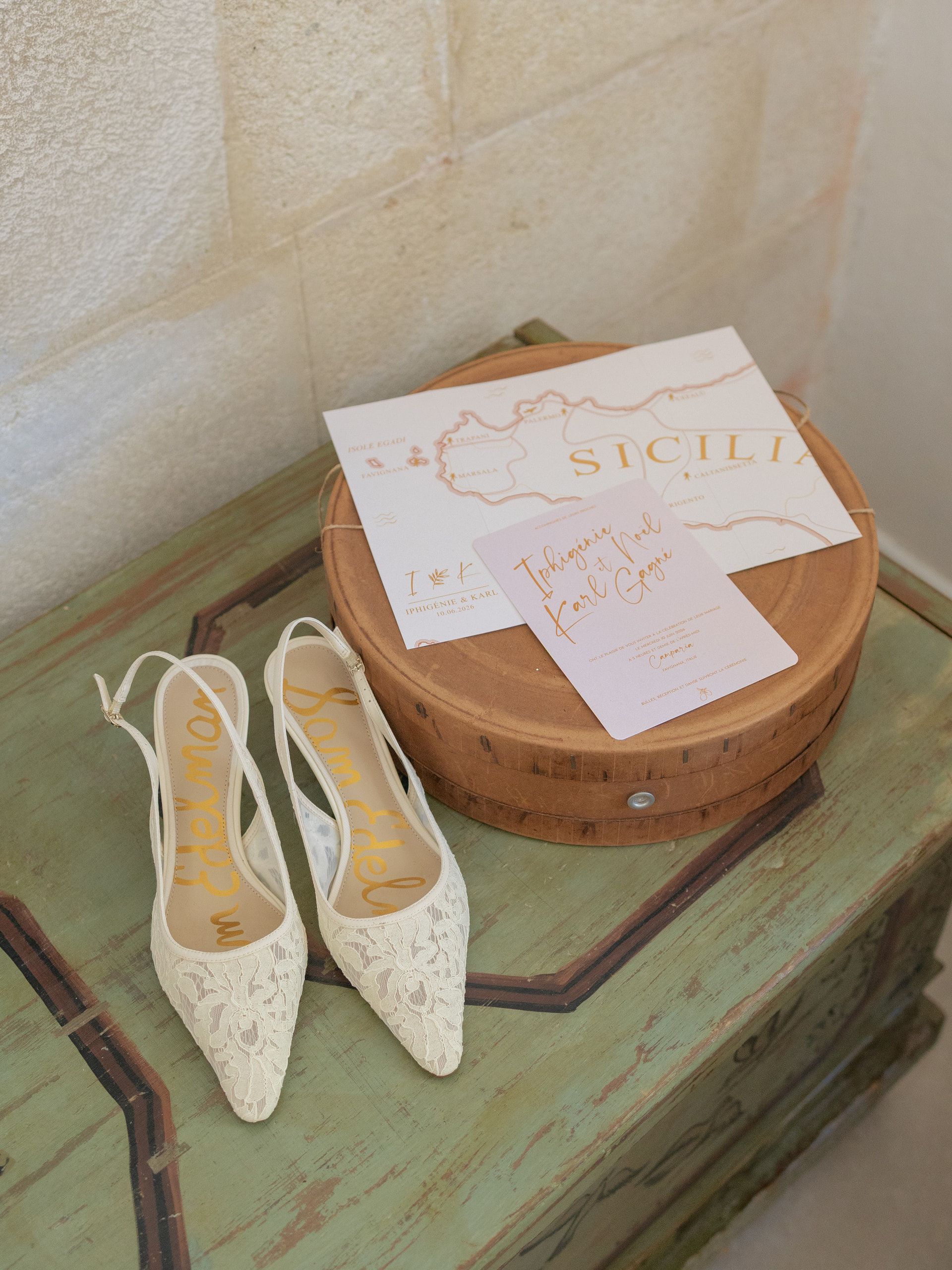 Le scarpe della sposa, in pizzo bianco, accanto a una scatola rotonda di legno con gli inviti, su un mobile verde