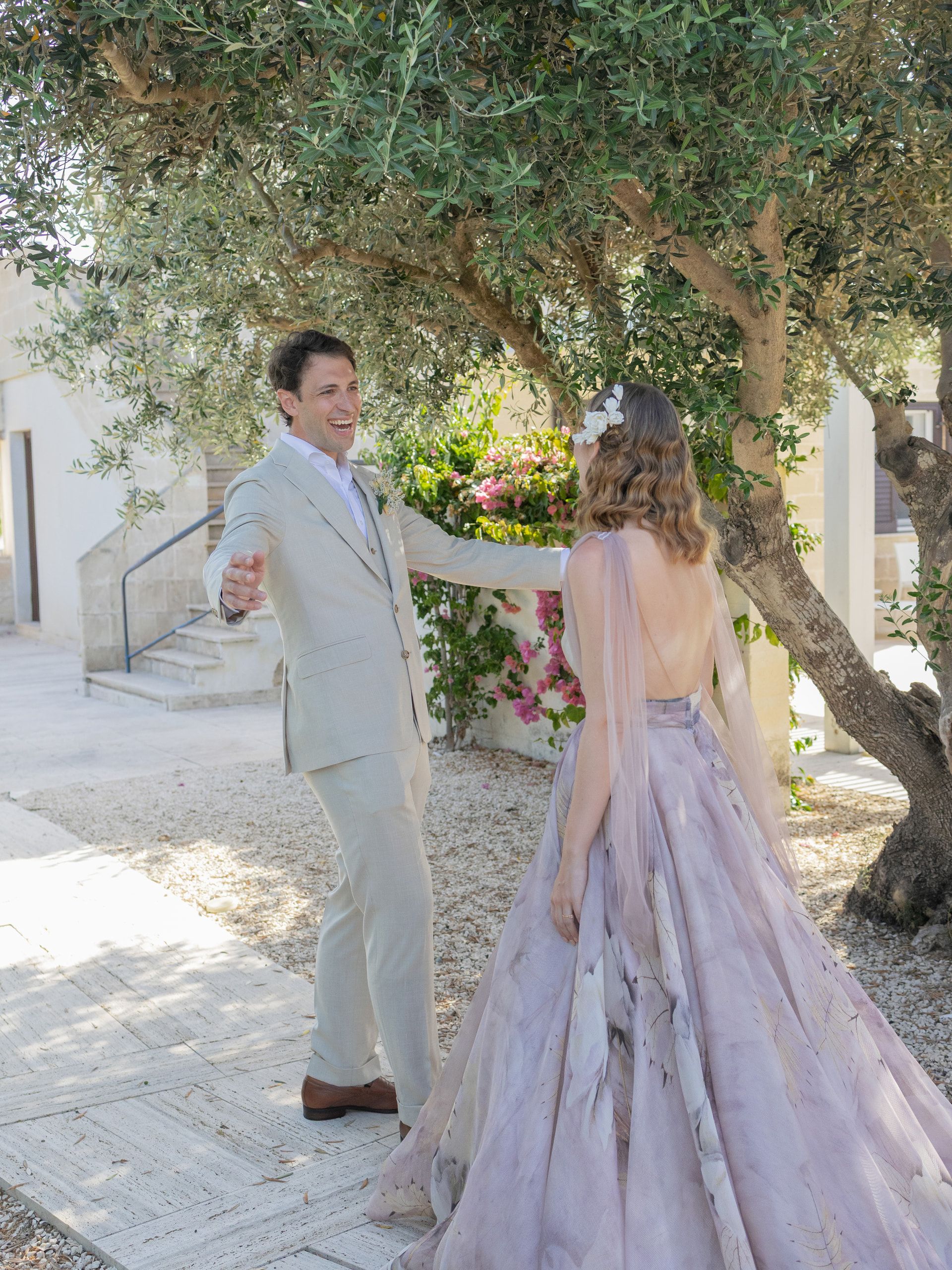 Lo sposo si volta a braccia aperte verso la sposa, ripresa di spalle, sotto un ulivo