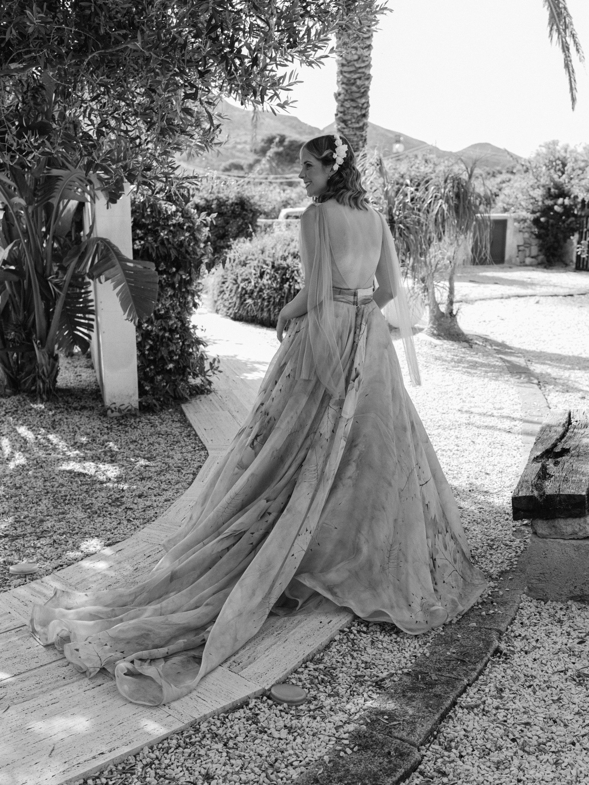 In bianco e nero, la sposa di spalle cammina su un vialetto di ghiaia fra le palme, con lo strascico dietro di sé