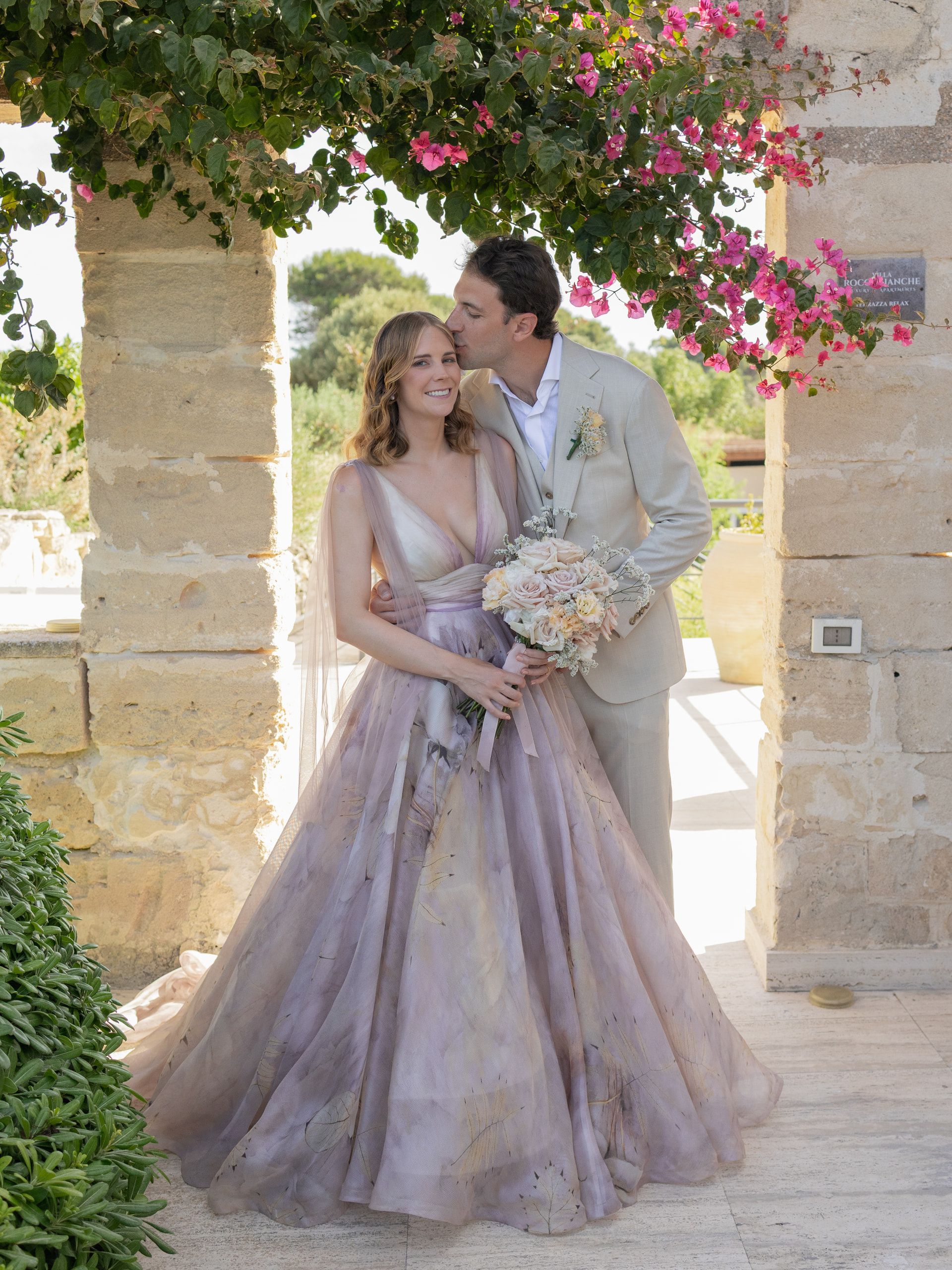 Lo sposo bacia la sposa sulla tempia sotto un arco di pietra coperto di bougainvillea