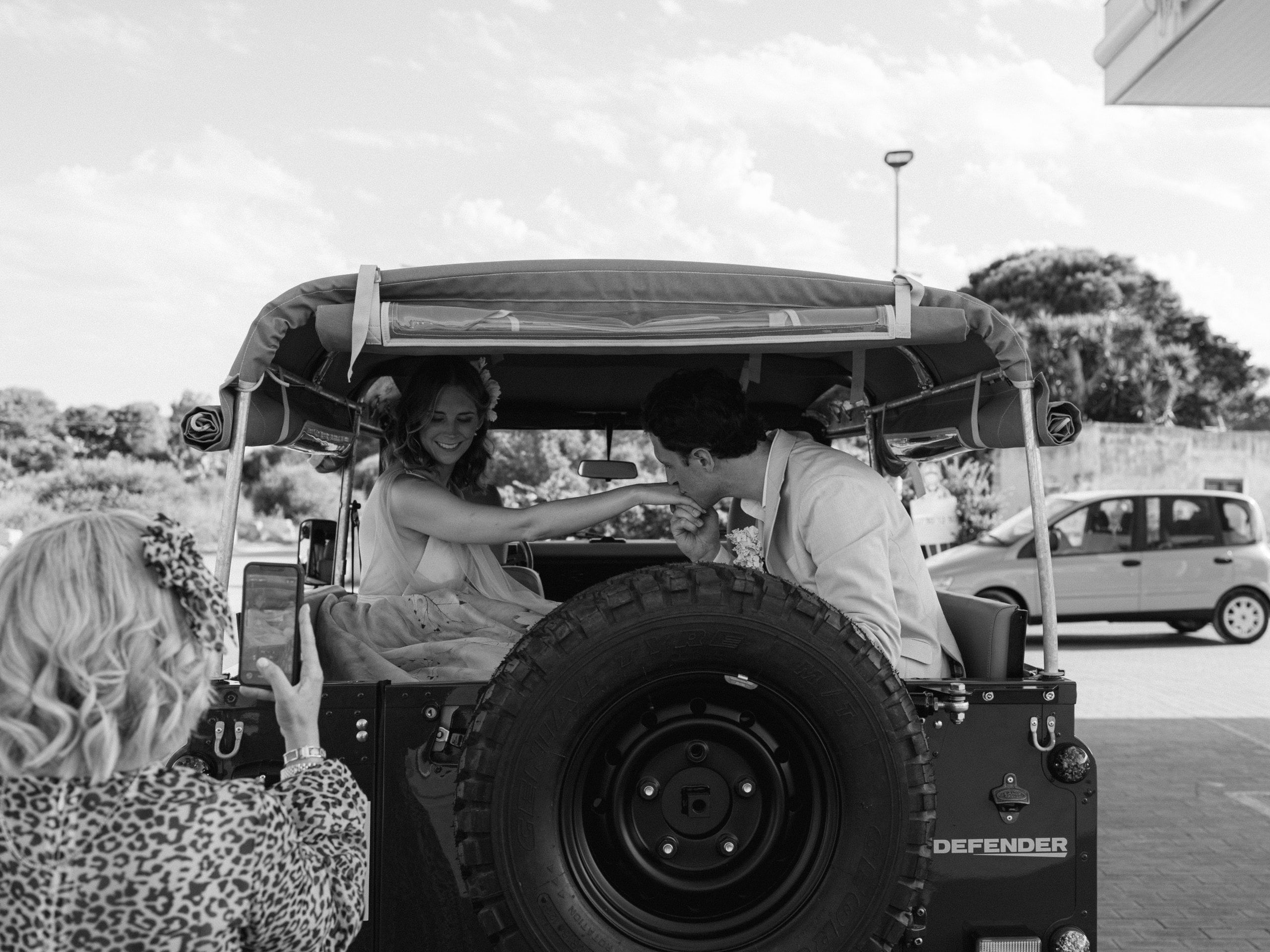 In bianco e nero, gli sposi seduti su una jeep scoperta; in primo piano un'invitata li fotografa con il telefono
