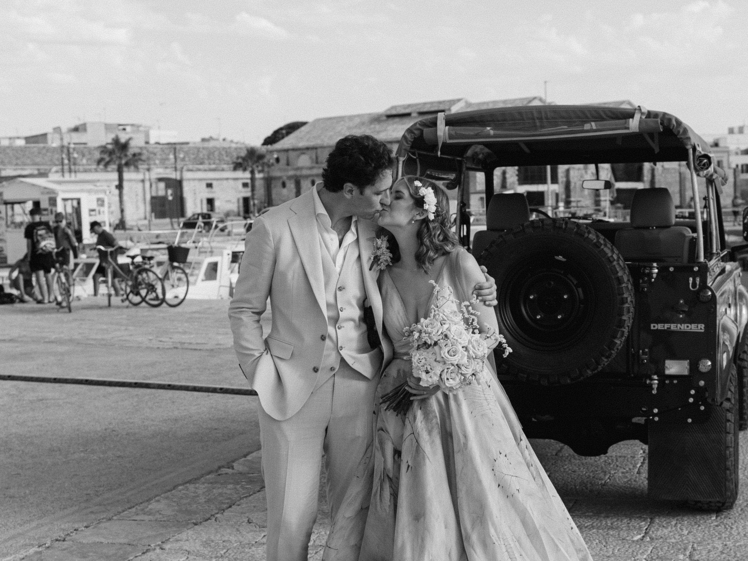 In bianco e nero, gli sposi si baciano accanto alla jeep, sulla banchina del porto