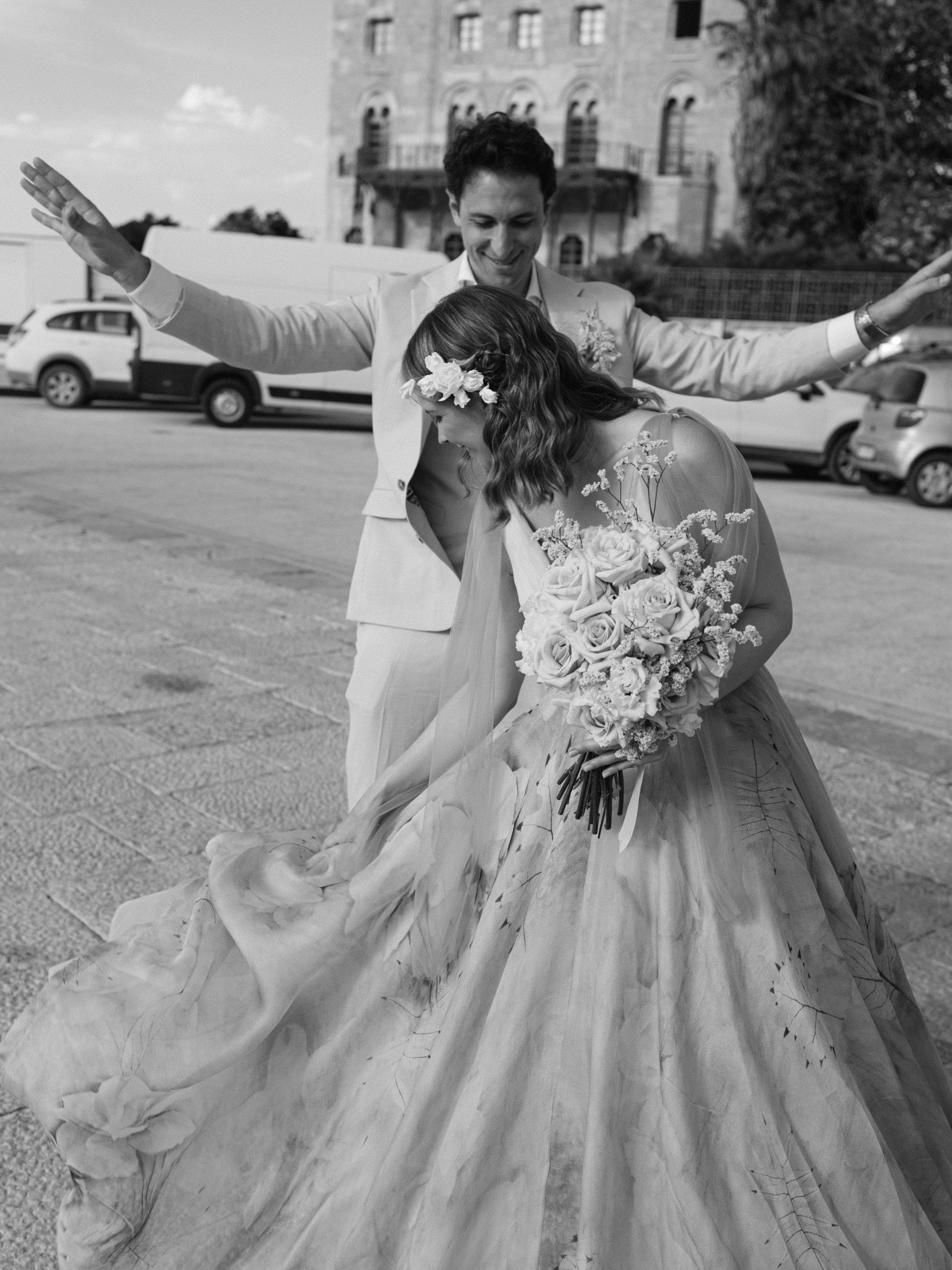 In bianco e nero, la sposa ride piegata sul bouquet mentre lo sposo, dietro di lei, alza le braccia in piazza