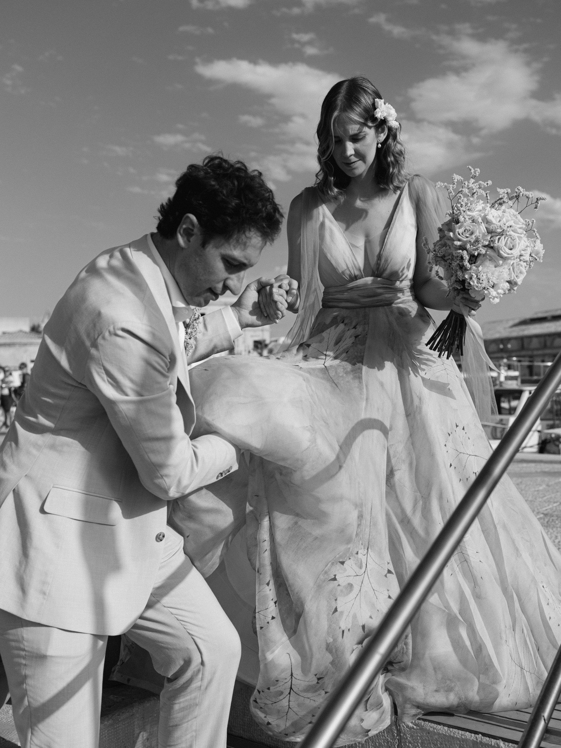 In bianco e nero, lo sposo solleva lo strascico della sposa che sale una scala con il bouquet in mano
