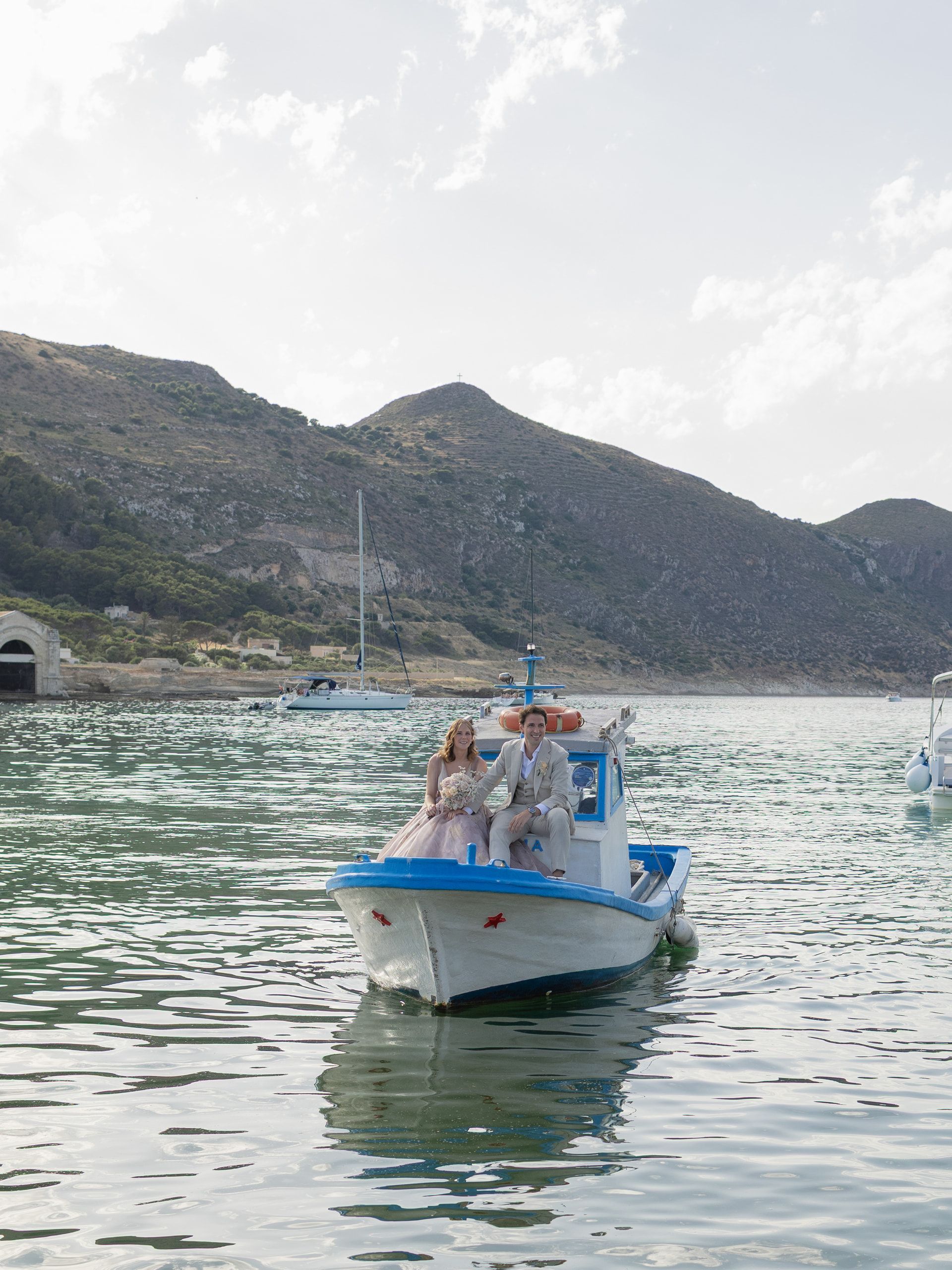 Gli sposi su una piccola barca bianca e azzurra nel mare calmo, con le colline dell'isola sullo sfondo