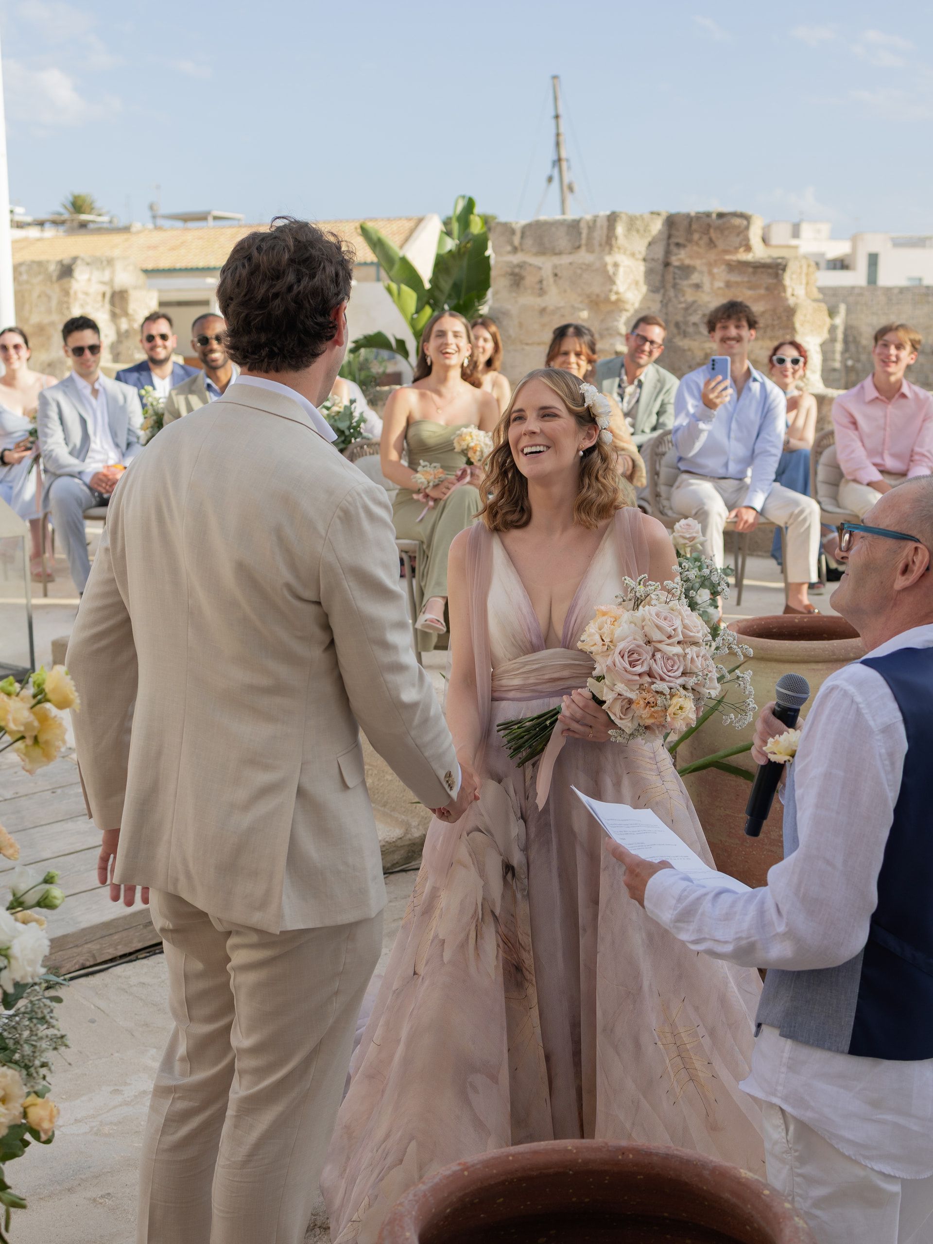 Durante la cerimonia all'aperto la sposa sorride allo sposo, davanti agli invitati seduti; il celebrante legge un foglio