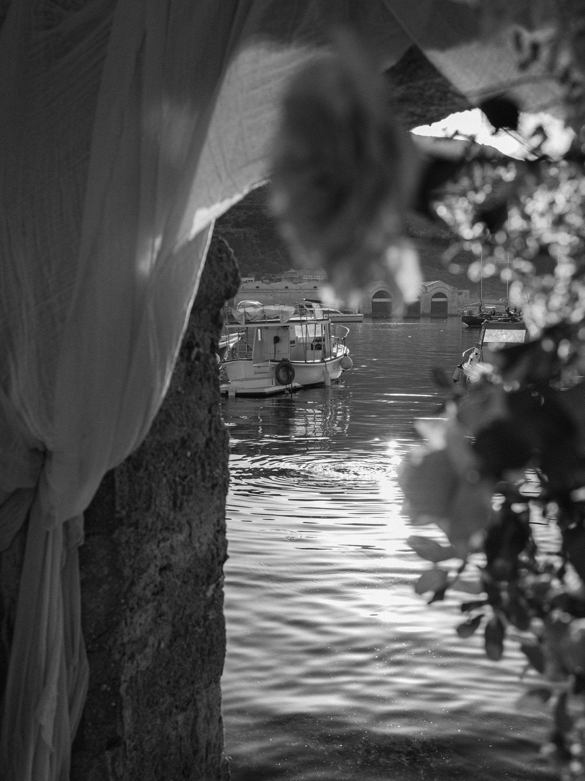 In bianco e nero, il mare del porto con le barche, visto fra un drappo e i fiori dell'arco