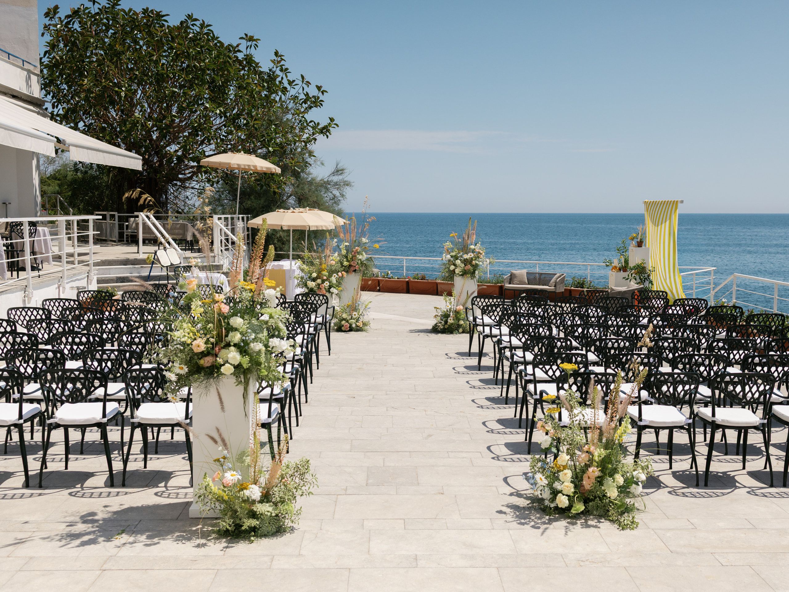 La terrazza sul mare pronta per la cerimonia: file di sedie nere, fiori lungo il corridoio e un drappo a righe gialle in fondo