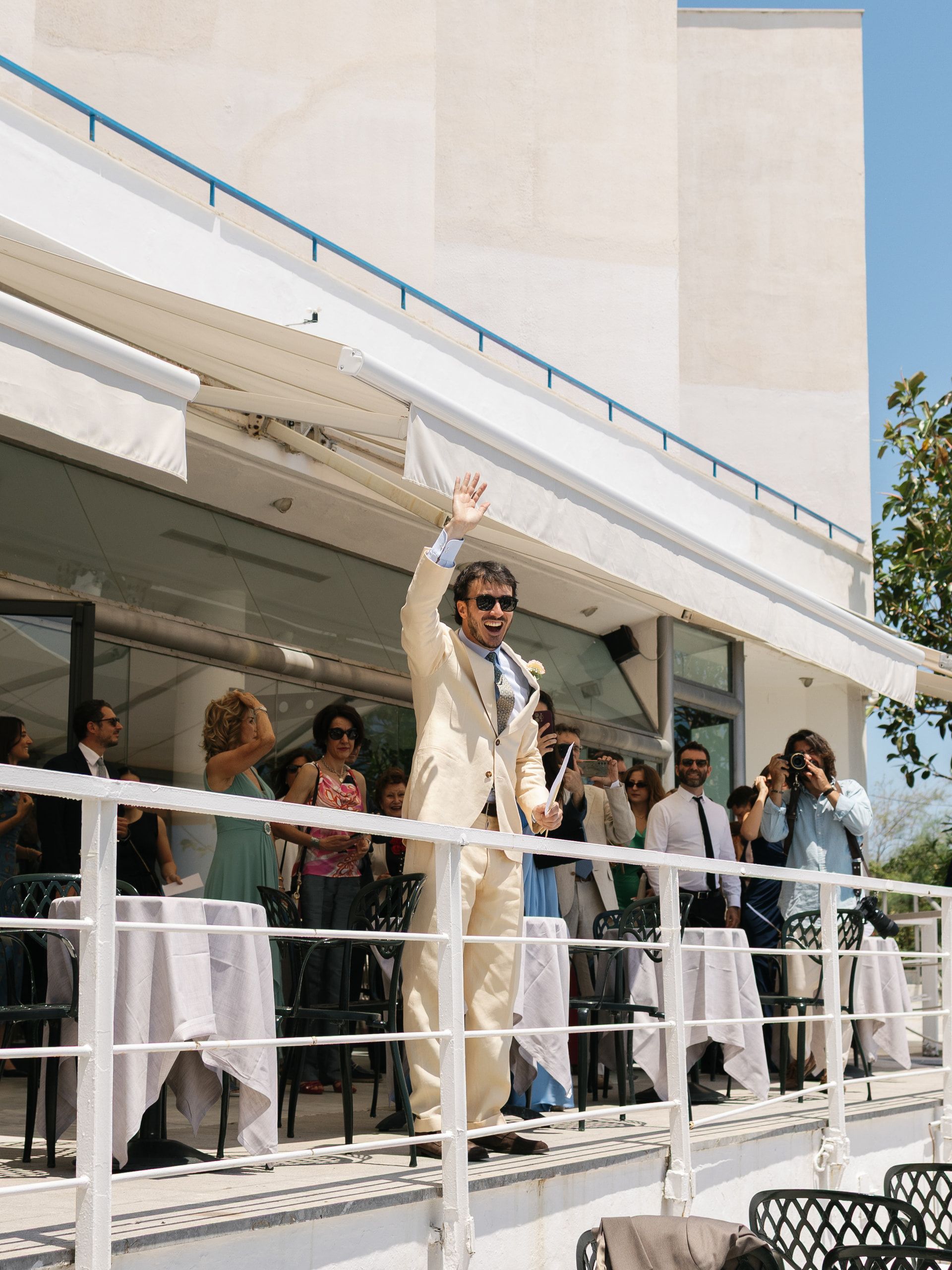 Lo sposo saluta con il braccio alzato da una terrazza, fra gli invitati sotto la tettoia