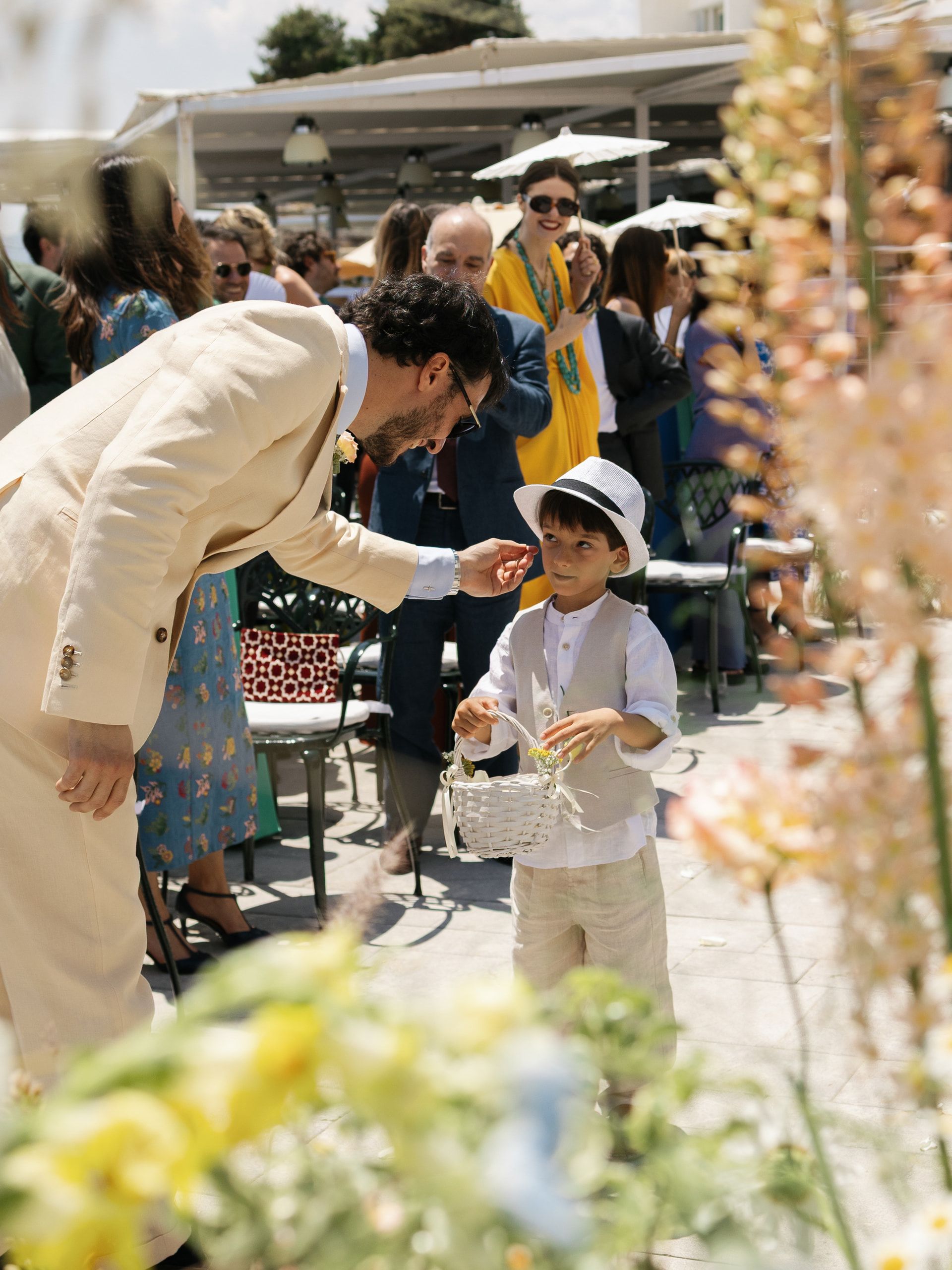 Lo sposo si china verso un bambino con un cappello bianco e un cestino, fra gli invitati