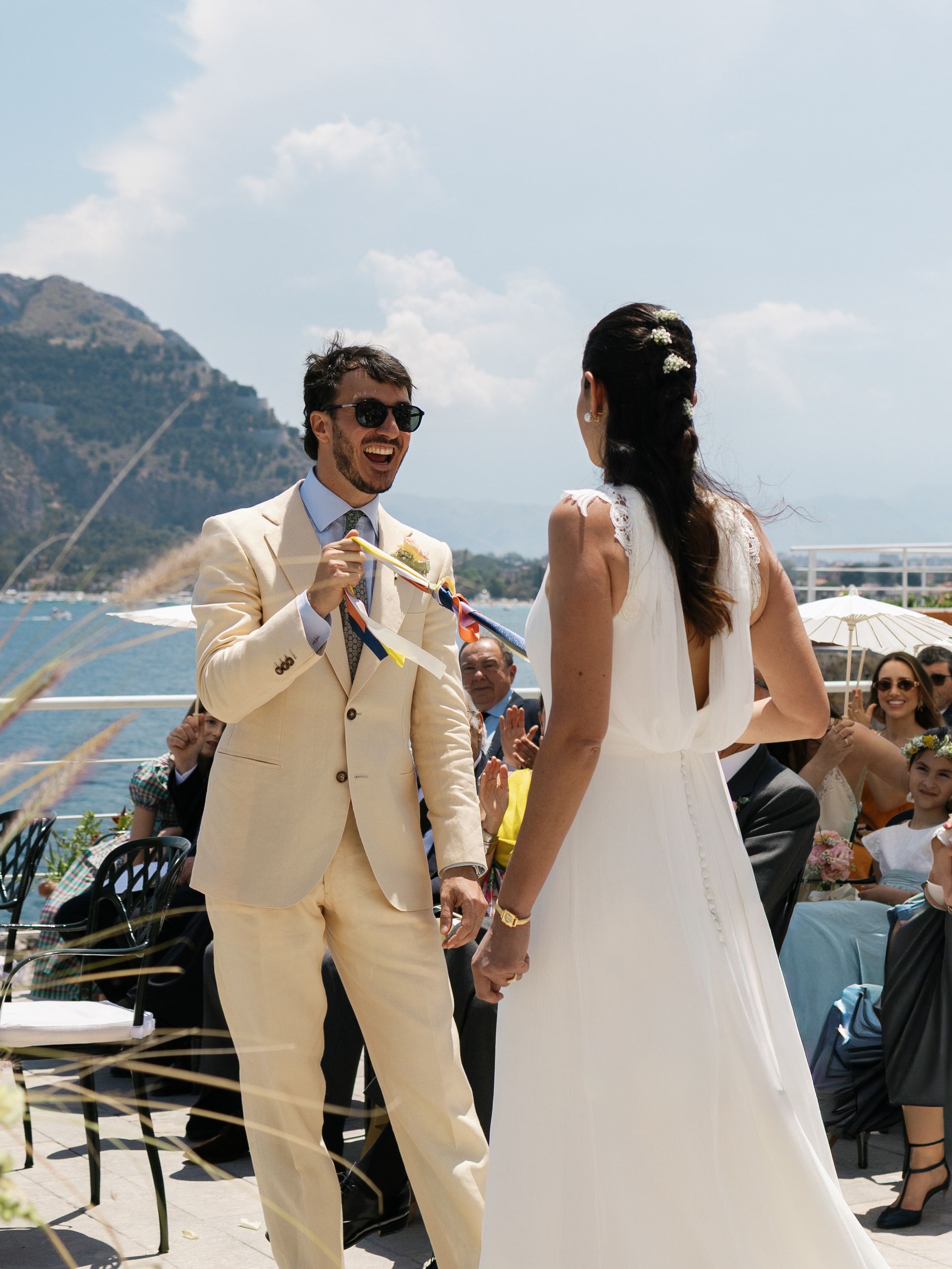 Lo sposo ride guardando la sposa, ripresa di spalle, durante la cerimonia sul mare
