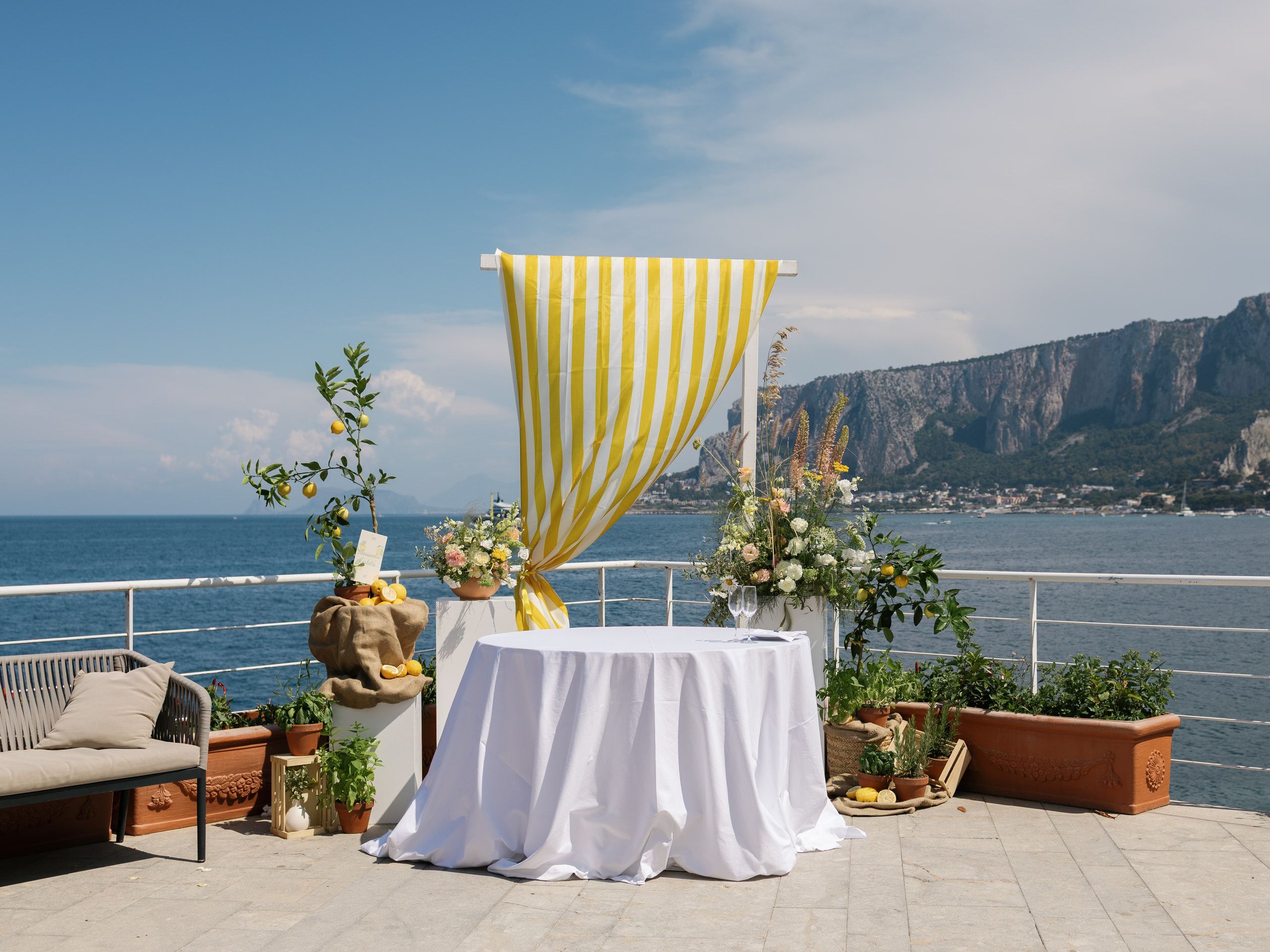 La terrazza ancora vuota: un tavolo con la tovaglia bianca sotto il drappo a righe gialle, il golfo e il promontorio sullo sfondo