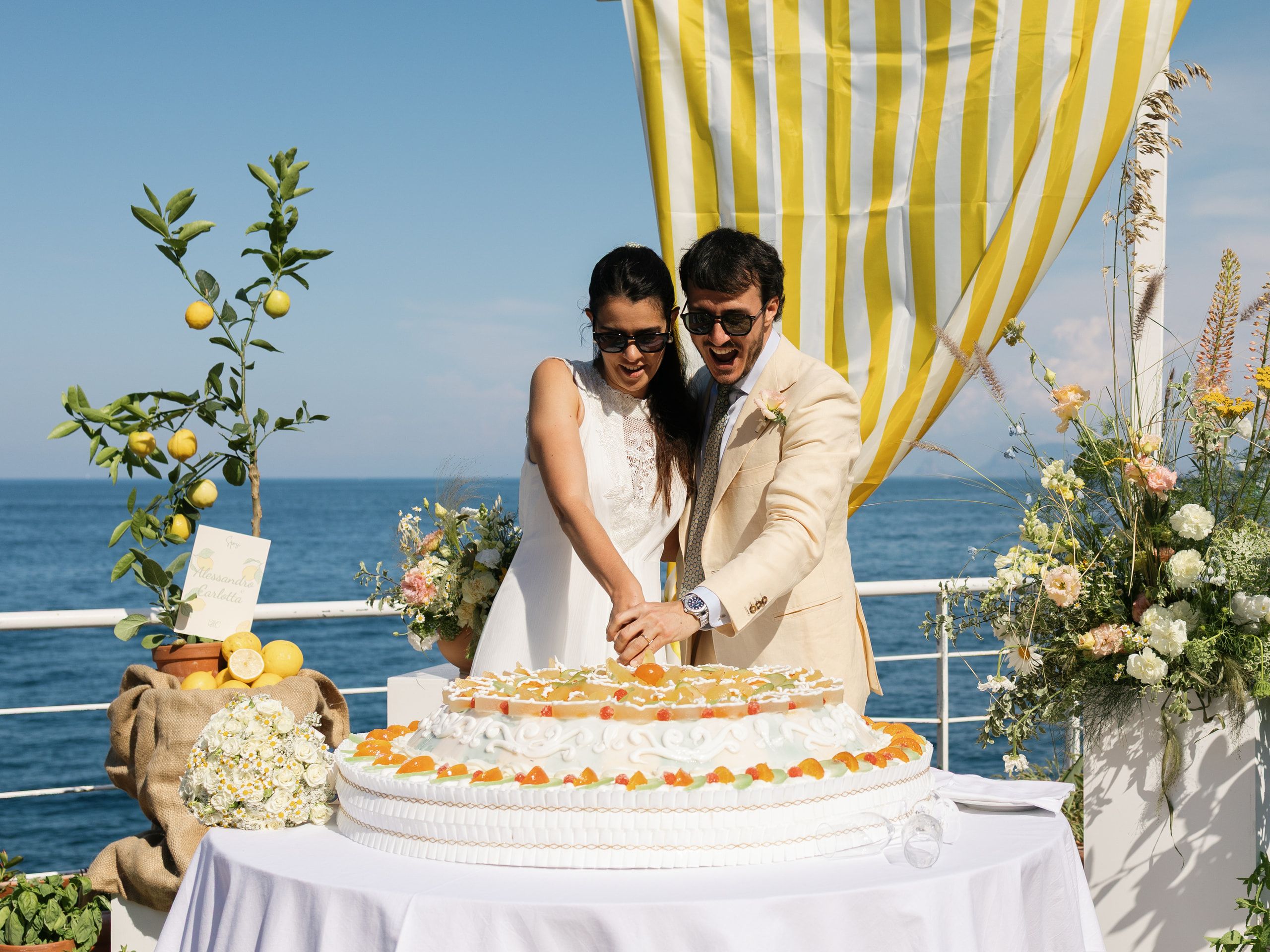 Gli sposi tagliano insieme la torta nuziale sotto il drappo a righe gialle, con il mare alle spalle