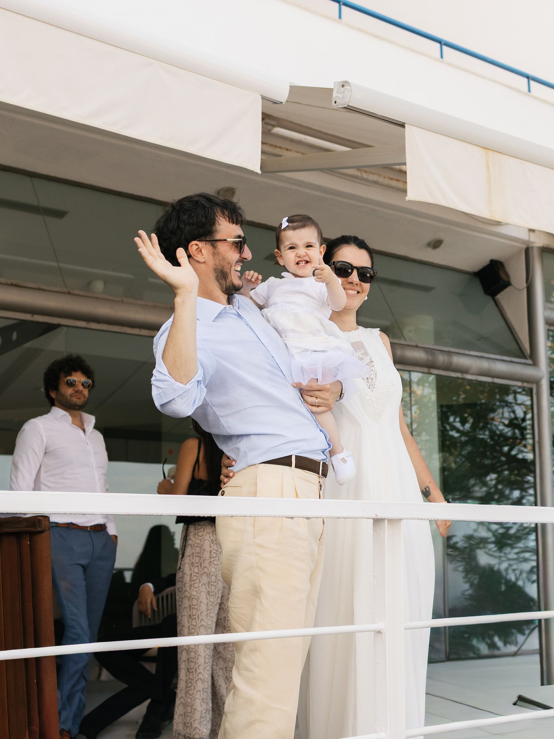 Un uomo con una bambina in braccio saluta da una terrazza, accanto alla sposa
